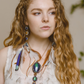 A model with curly hair looking sideways, showcasing the elegance of the Azure Key Amulet from the You Are The Key Collection. This handcrafted piece features Malachite, Azurite, Clear Quartz, Lapis, and Gold filled silver—a symbol of enlightened leadership. Paired with a 1.5mm snake chain, 20 inches. Comes with a custom silk bag for safekeeping. Capture the essence of the Azure Key—a unique blend of gemstones for personal transformation.